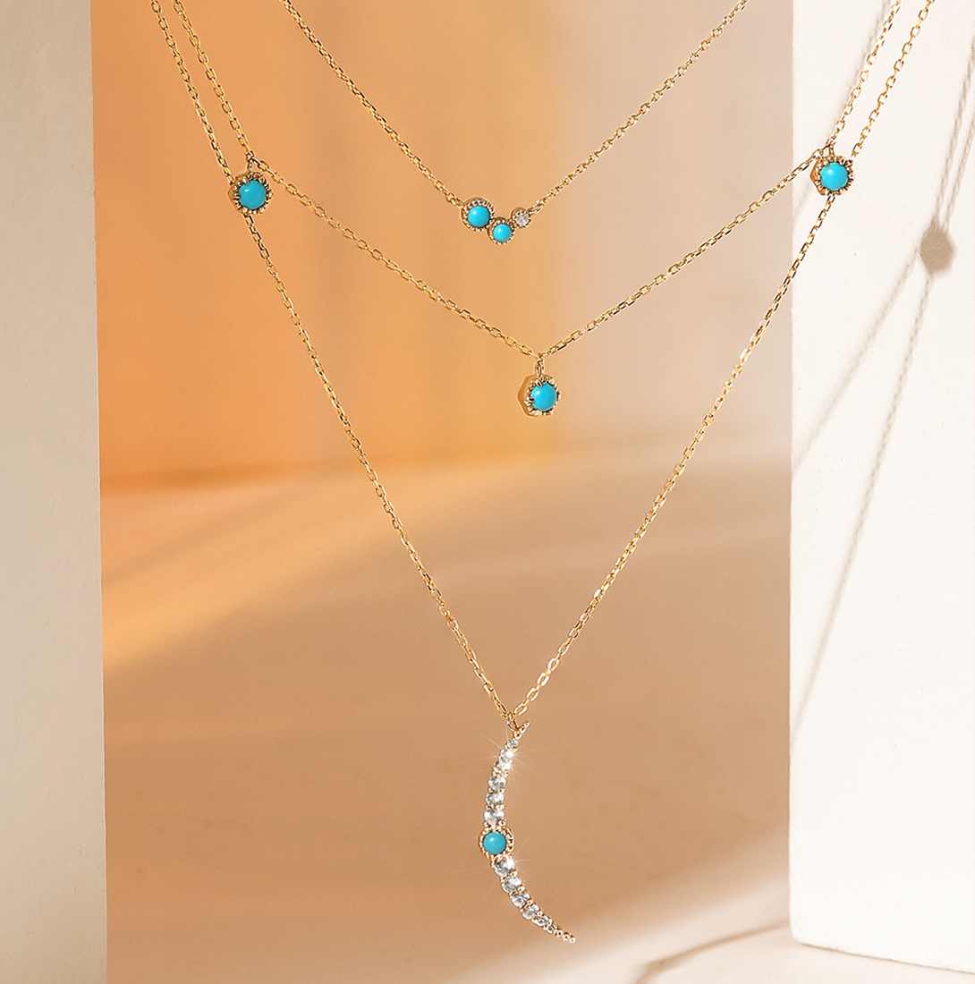 Aurelie Gi layered gold necklaces featuring turquoise gemstones and a crescent moon pendant accented with sparkling white stones, displayed in warm golden light.