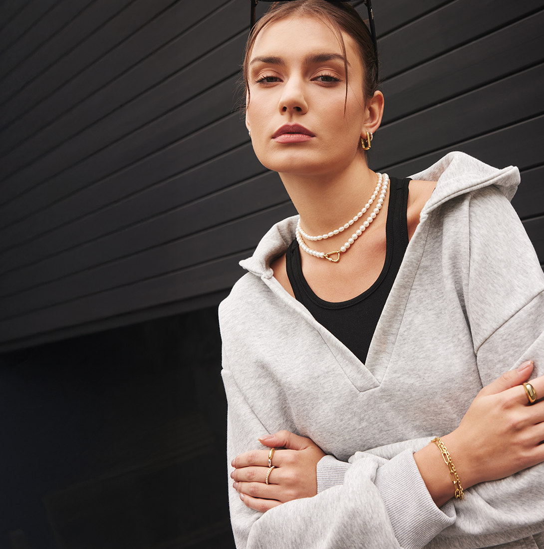 Stylish woman wearing Ania Haie jewelry, including layered pearl necklaces with a gold heart clasp, gold hoop earrings, and gold rings and bracelet. She’s dressed in a light gray zip sweatshirt over a black tank, posing confidently against a modern dark background.