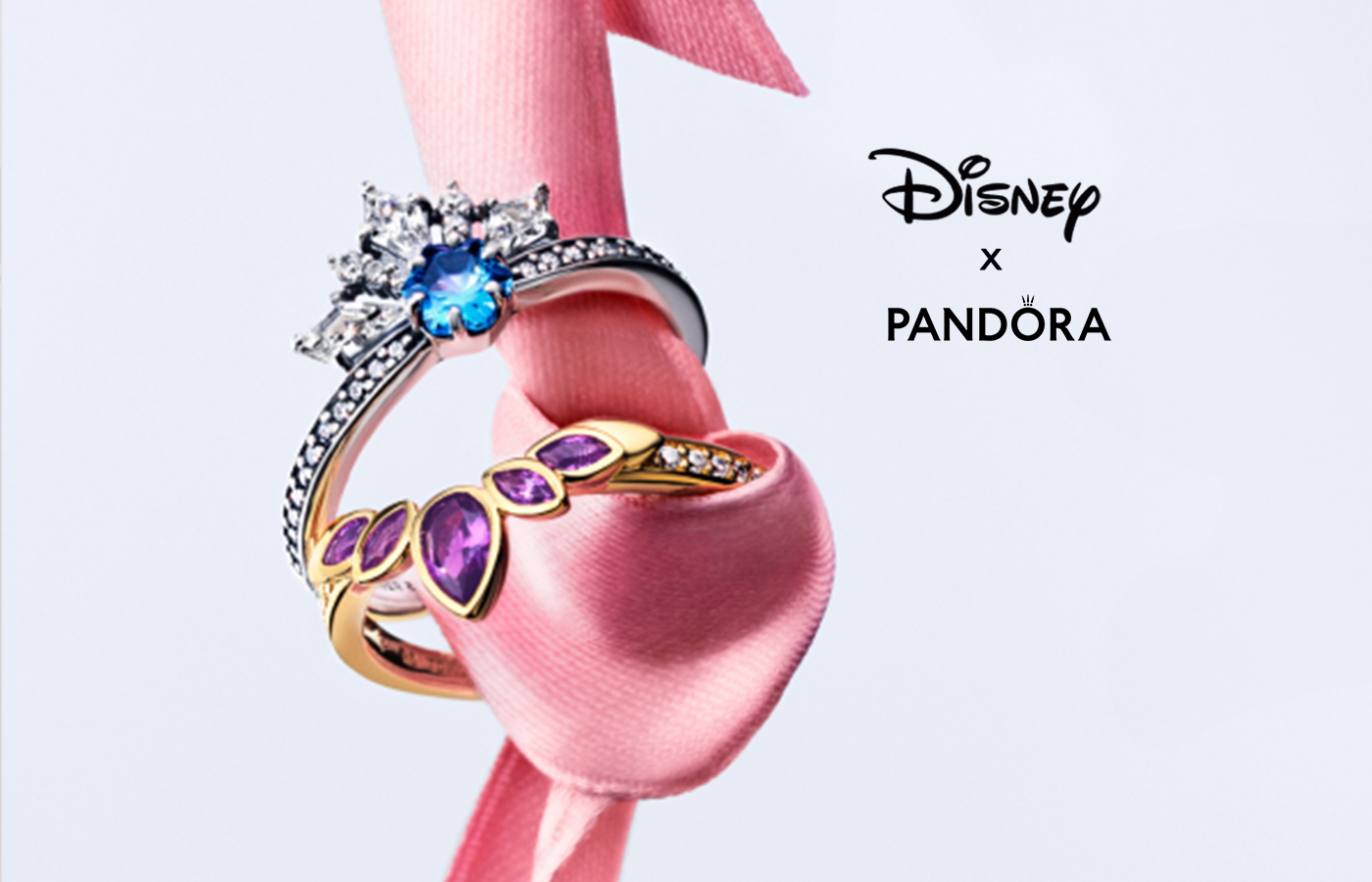 Disney x Pandora rings featuring a silver snowflake-inspired ring with blue gemstone and a gold ring with purple marquise stones, styled on pink satin ribbon.