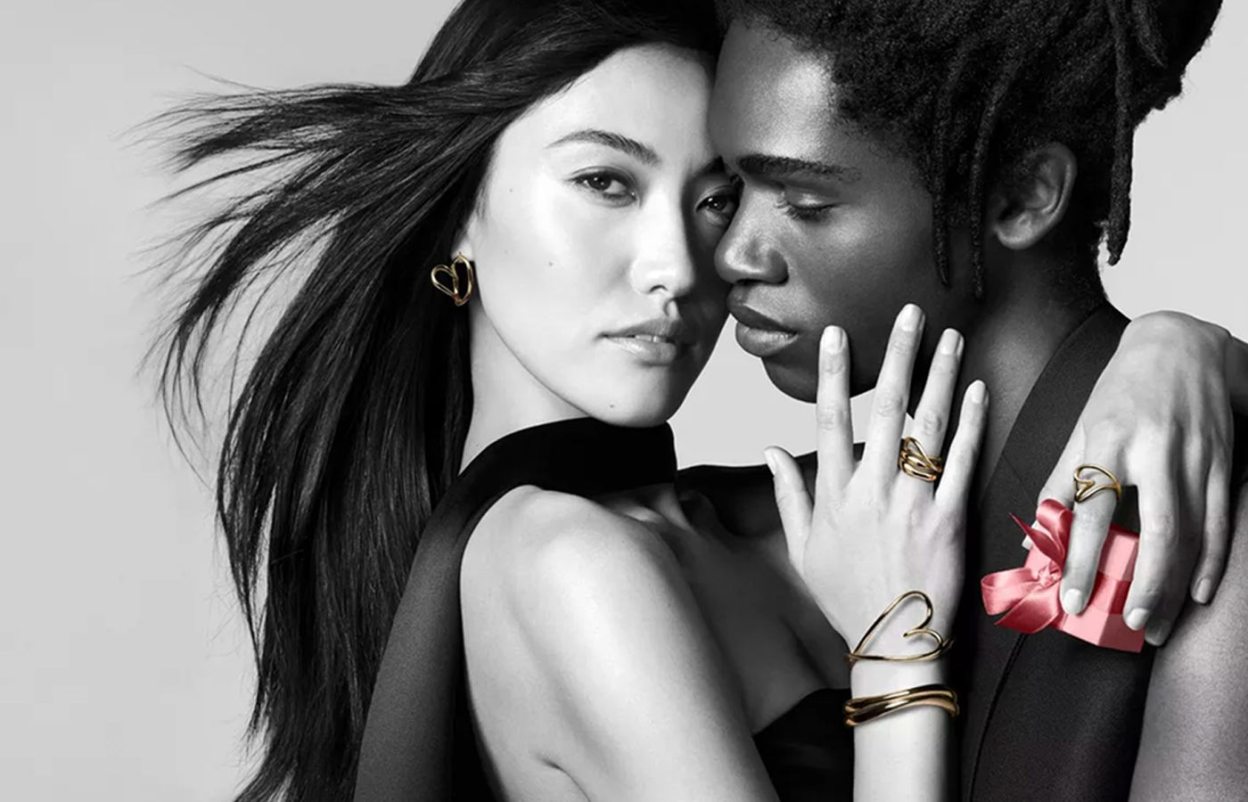 Stylish couple embracing while wearing sculptural gold jewelry from the Pandora Essence Collection, including heart-shaped earrings, rings, and bracelets — with a pink gift box accent for a bold, modern expression of love and connection.